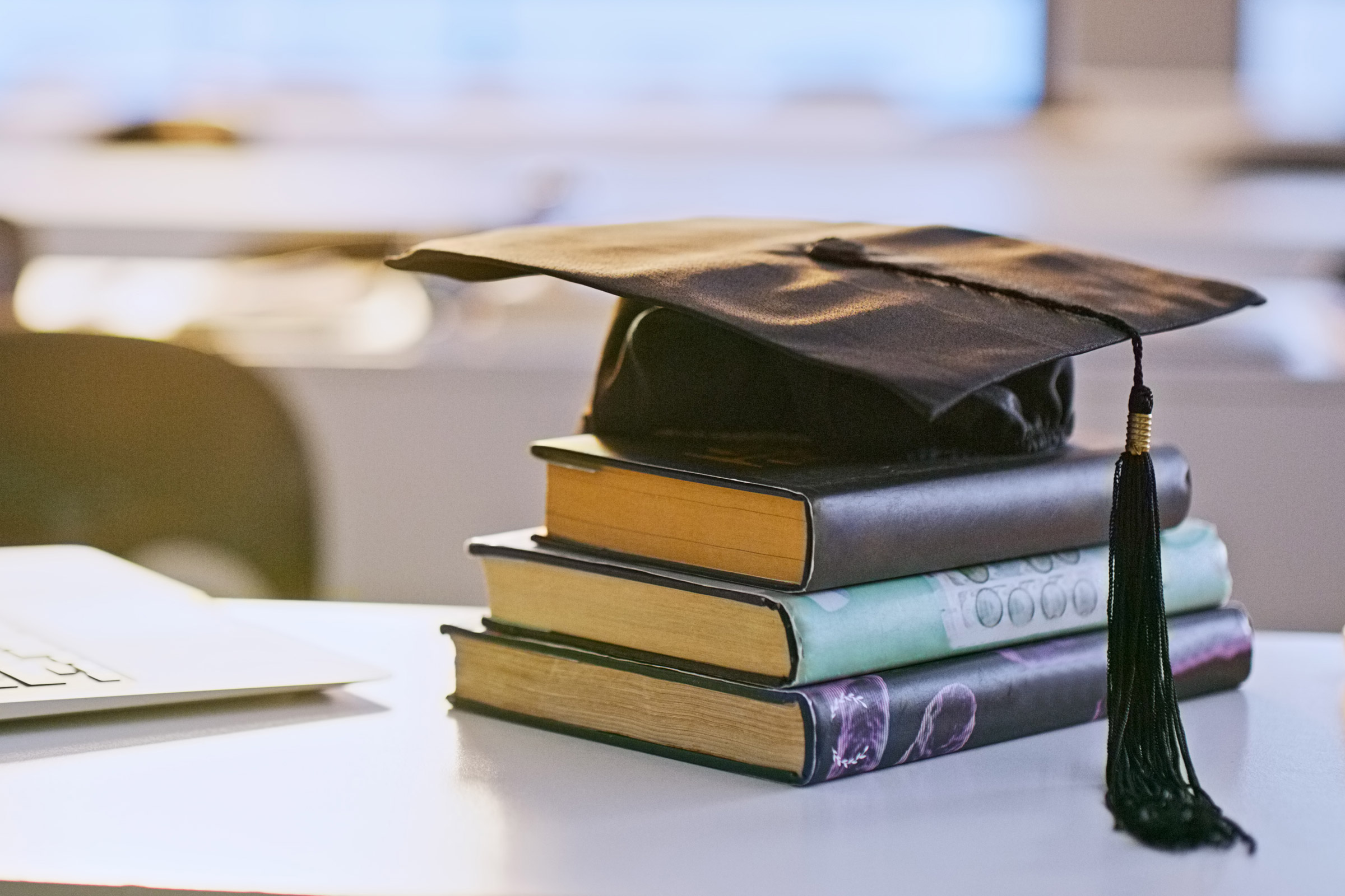Graduation cap on stacked books beside laptop
